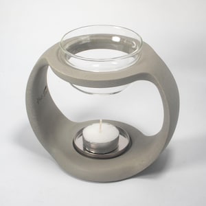 May include: A gray, sculptural candle holder with a clear glass bowl at the top and a small tea light candle at the bottom. The holder has a modern, curved design with a circular opening.