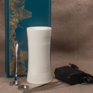 Op de afbeelding: Witte keramische aromatherapie-diffuser met een cilindrische vorm en horizontale ribbels. Accessoires: zilveren lepel, pincet en zwarte voedingsadapter. Een blauwgroene doos met gouden decoratieve patronen op de achtergrond.