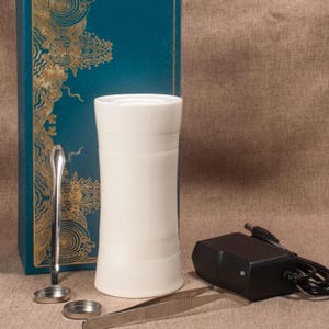 Può includere: Diffusore per aromaterapia in ceramica bianca, di forma cilindrica con scanalature orizzontali. Accessori: cucchiaio d'argento, pinzette e adattatore di alimentazione nero. Su una scatola turchese con motivi dorati.