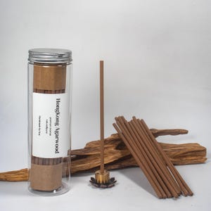 May include: A clear glass tube with a silver lid contains incense sticks, labeled "Hongkong Agarwood." Several incense sticks are arranged on a wooden surface, with one burning in a decorative holder. The incense is a light brown color.
