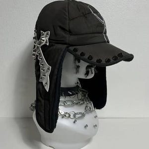May include: Black trapper hat with a quilted design, featuring a curved brim with black studs. The hat has ear flaps and is adorned with a silver decorative element. A silver chain necklace is also visible.
