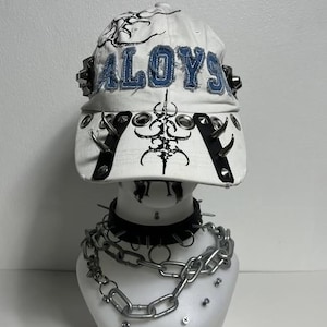 Op de afbeelding: Witte baseballcap met het woord "ALOY" in blauwe letters. De cap heeft zwarte spikes en decoratieve metalen accenten. Een zwarte choker met spikes en een zilveren ketting zijn ook zichtbaar.
