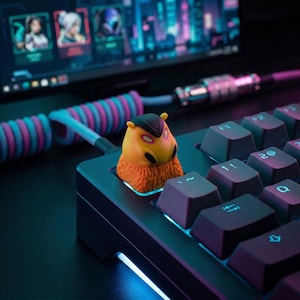 Puede incluir: Un teclado mecánico negro con una tecla personalizada con un diseño de criatura amarilla y naranja. El teclado tiene un cable en espiral con detalles en rosa y azul, y un monitor de ordenador es visible en el fondo.