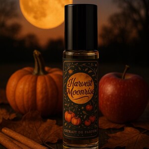 Op de afbeelding: Een parfumflesje met een zwarte dop en een etiket met de tekst "Harvest Moonrise" met pompoenillustraties. De fles is omgeven door een pompoen, een appel, kaneelstokjes en herfstbladeren. Op de achtergrond is een grote, oranje maan te zien.