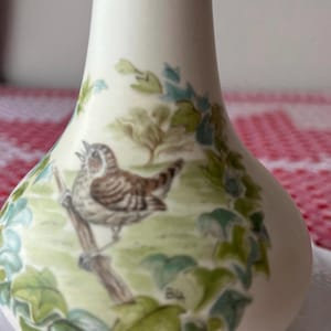 May include: A cream-coloured ceramic vase featuring a detailed illustration of a small brown bird perched on a branch. The bird is surrounded by green foliage and ivy leaves. The artwork includes the initials BL in the lower right corner of the design.