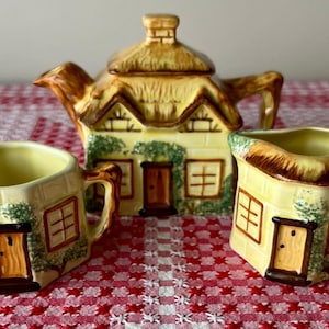 May include: A delightful cottage-style tea set, including a teapot and two cups. The set is painted in a pale yellow with brown detailing, depicting windows, doors, and thatched roofs. The set is displayed on a red and white checked tablecloth.