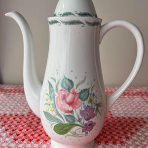 May include: White ceramic coffee pot with a floral design featuring pink roses, purple and blue flowers, and green leaves. The pot has a curved spout, a handle, and a lid with a green leaf pattern. Small gray dots are scattered across the white surface.