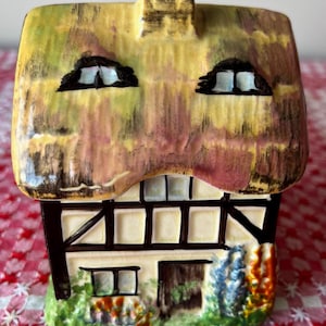 May include: A ceramic cottage figurine featuring a yellow thatched roof with a chimney, dark brown timber framing on cream walls, two upper windows, a central door, and painted green foliage with colourful flowers at the base of the structure.