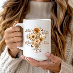 May include: A white ceramic mug featuring a central cross design composed of orange and cream-colored flowers and small butterflies. The text reads: in the WAITING, in the HEALING, in the HURTING, in the SEARCHING, as there was on JESUS.