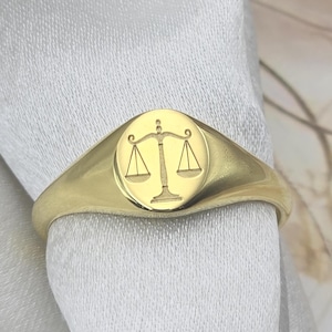 Peut inclure: Bague chevalière dorée avec une gravure de balance, symbole de la justice. La bague a une finition polie et une face ovale. La bague est présentée sur un fond de tissu blanc. La bague est en or.