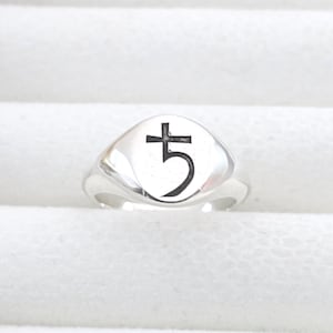 May include: A silver signet ring with a polished finish. The ring features a black symbol on the oval face. The symbol is a cross with a curved line extending from the bottom. The ring is set against a white background.