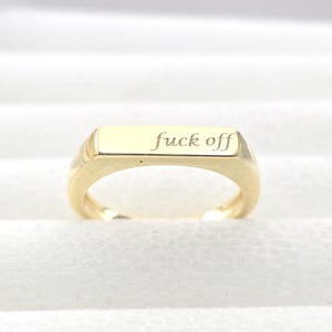 Puede incluir: Un anillo dorado con una parte superior rectangular. El anillo tiene la frase "fuck off" grabada en la parte superior. El anillo está sobre un fondo blanco.