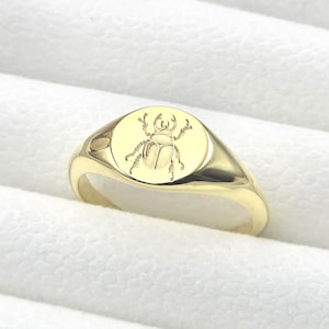 Könnte beinhalten: Ein goldener Siegelring mit einer detaillierten Gravur eines Käfers auf der kreisförmigen Oberfläche. Der Ring hat eine glatte, polierte Oberfläche und ein klassisches Design. Der Ring wird auf einer weißen, strukturierten Oberfläche präsentiert.