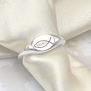 Puede incluir: Un anillo plateado con una cara ovalada que presenta la palabra griega "ΙΧΘΥΣ" y un símbolo de pez. El anillo se muestra sobre un tejido blanco y sedoso, resaltando su acabado pulido y su diseño.