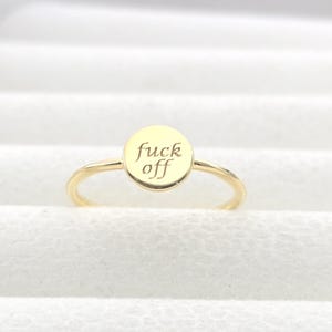 Puede incluir: Anillo dorado con una cara redonda. La cara del anillo tiene las palabras "fuck off" grabadas en una fuente cursiva. El anillo está sobre un fondo blanco.