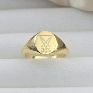 Puede incluir: Un anillo de sello dorado con una cara ovalada. El anillo presenta un símbolo grabado con un triángulo y líneas que se cruzan, y el número 302 debajo. El anillo está sobre un fondo blanco.