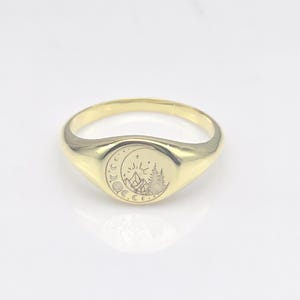 Peut inclure: Une bague chevalière dorée avec un motif gravé de montagne et de lune. La bague présente une finition polie et une face ovale arrondie. Le motif gravé comprend un croissant de lune, des étoiles et une forêt.