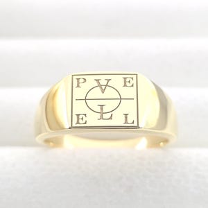 May include: A gold signet ring with a rectangular face. The face of the ring has the letters P, V, E, and L, along with a circle and a horizontal line. The ring is made of polished gold.