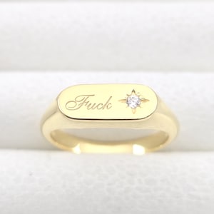 Op de afbeelding: Een gouden zegelring met het woord "Fuck" gegraveerd op het rechthoekige vlak. Een klein, stervormig ontwerp met een heldere edelsteen bevindt zich rechts van het woord. De ring staat tegen een witte achtergrond.