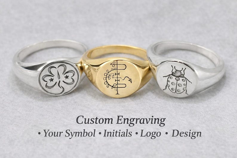 May include: Three signet rings with custom engravings. One silver ring features a clover design, a gold ring has a symbol, and another silver ring displays a ladybug. The text "Custom Engraving" and options are below the rings.