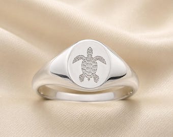May include: A polished silver signet ring featuring an oval face engraved with a detailed sea turtle design. The ring has a smooth, rounded band and a reflective metallic finish, showcasing a clear, centered illustration of the turtle.
