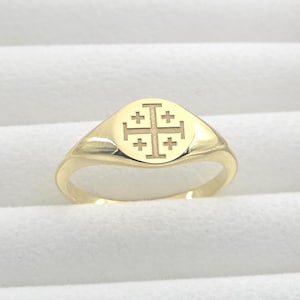 以下が含まれることがあります： 丸みを帯びた顔の金色の指輪。 指輪には、キリスト教の象徴であるエルサレム十字のデザインが施されています。 指輪は白い背景に配置され、磨かれた表面が強調されています。 指輪のバンドは滑らかで丸みを帯びています。