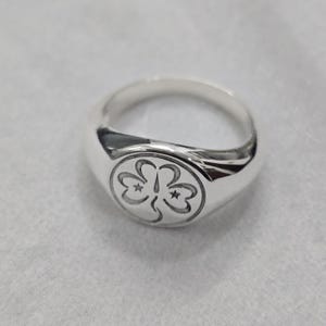 May include: A silver signet ring with a rounded face. The ring features a detailed emblem of three stylized clover-like shapes, each with a star. The ring is made of polished metal and sits on a light gray surface.