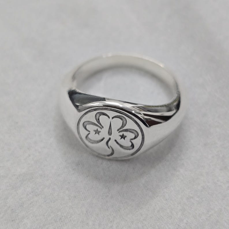 May include: A silver signet ring with a rounded face. The ring features a detailed emblem of three stylized clover-like shapes, each with a star. The ring is made of polished metal and sits on a light gray surface.