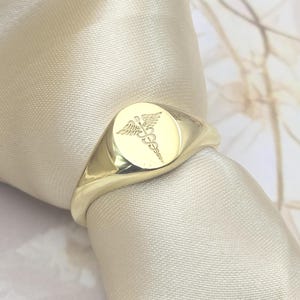 May include: A gold signet ring with a round face featuring the caduceus symbol. The ring is set against a cream-colored background, highlighting its polished, reflective surface. The ring's design is simple and elegant.