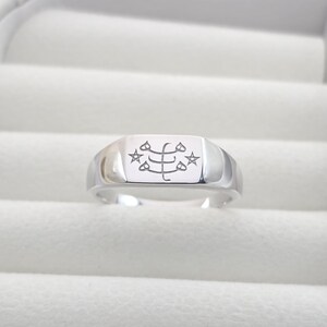 May include: A silver ring with a rectangular face featuring an engraved design with stars and symbols. The ring has a polished finish and a classic, minimalist style, suitable for everyday wear.