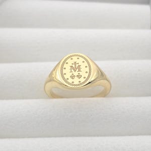 Puede incluir: Un anillo de sello dorado con una cara ovalada. La cara presenta un diseño grabado con una cruz, la letra "M", estrellas y corazones. El anillo está hecho de oro pulido y tiene una banda lisa y redondeada.