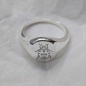 May include: A polished silver ring with a rounded face. The ring features a detailed engraving of a ladybug with dotted wings. The ring is a signet style, with a smooth, reflective surface.