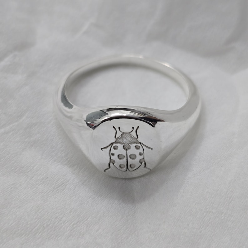May include: A polished silver ring with a rounded face. The ring features a detailed engraving of a ladybug with dotted wings. The ring is a signet style, with a smooth, reflective surface.