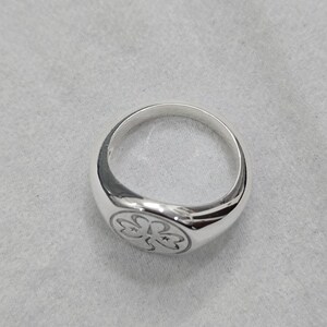 May include: A polished silver ring with a rounded band and a flat, circular face. The face of the ring features a detailed, engraved floral design. The ring is set against a neutral, textured background.