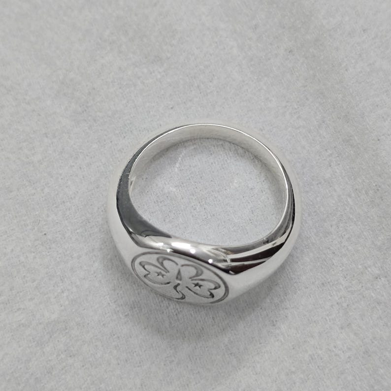 May include: A polished silver ring with a rounded band and a flat, circular face. The face of the ring features a detailed, engraved floral design. The ring is set against a neutral, textured background.