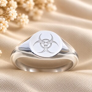 May include: A polished silver-tone signet ring featuring a circular face engraved with a biohazard symbol. The ring has a smooth, reflective metallic band and a flat, round top surface that displays the etched hazard icon clearly.