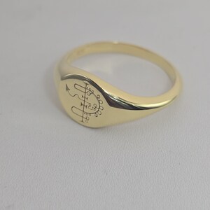 May include: A gold signet ring with a polished finish. The ring features an engraved symbol on the oval face. The band is smooth and rounded. The ring is displayed on a neutral surface.