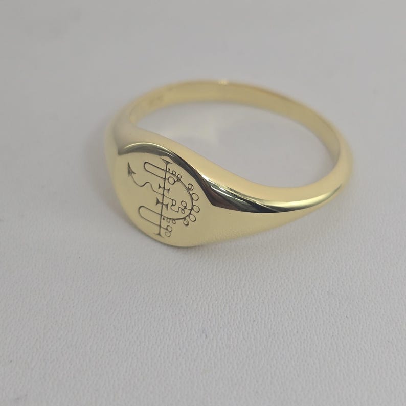 May include: A gold signet ring with a polished finish. The ring features an engraved symbol on the oval face. The band is smooth and rounded. The ring is displayed on a neutral surface.