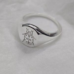 May include: A silver signet ring with a detailed engraving of a ladybug on the face. The ring has a smooth, polished finish and a rounded band. The ladybug design features spots and delicate details, creating a nature-inspired jewelry piece.