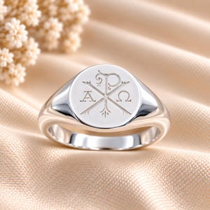 Puede incluir: Un anillo de sello de plata pulida con una cara circular grabada con el símbolo Chi-Rho, flanqueado por las letras griegas Alfa y Omega. El anillo presenta una banda metálica lisa y reflectante con una superficie superior plana y circular.
