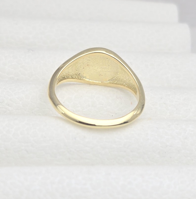 May include: A gold-colored ring with a smooth, oval-shaped face. The ring has a simple, classic design, and the band is rounded. The ring is likely made of gold or a gold-colored metal. The ring is a piece of jewelry.