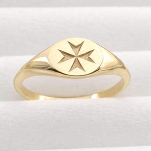 以下が含まれることがあります： 丸い顔にマルタ十字のデザインが施されたゴールドカラーのリング。 リングは光沢のある仕上げとシンプルなバンドが特徴です。 十字架はリングの顔にエッチングされています。 リングは白い背景に配置されています。