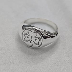 May include: A silver signet ring with a rounded face. The face features a detailed, engraved design of three stylized floral shapes, each with a star in the center. The ring is made of polished metal.