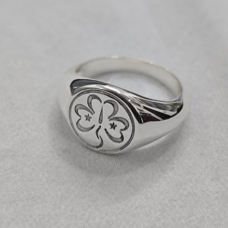 May include: A silver signet ring with a rounded face. The face features a detailed, engraved design of three stylized floral shapes, each with a star in the center. The ring is made of polished metal.