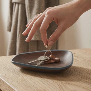 May include: A dark brown wooden tray with a smooth, rounded triangular shape and a dark grey rim. The tray contains a set of metal keys attached to a small brown leather tag. The text "Le geste en passant la porte. Clés posées en rentrant, retrouvées en partant." is visible at the top.