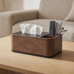Puede incluir: Un soporte rectangular para caja de pañuelos de madera oscura con un panel superior negro y un compartimento lateral integrado. El compartimento contiene un mando a distancia negro, uno blanco y unas gafas de montura marrón. Sobresalen pañuelos blancos.