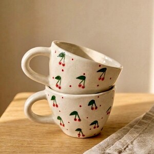 Puede incluir: Dos tazas de cerámica apiladas con una base de color crema, con un patrón pintado a mano de pequeñas cerezas rojas con hojas verdes y puntos rojos dispersos. Cada taza tiene un cuerpo redondeado y un asa curva con un acabado brillante.