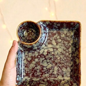 Puede incluir: Un plato de servicio cuadrado de cerámica con un cuenco circular integrado para salsas. La pieza presenta un esmalte brillante de color marrón oscuro con un patrón cristalino moteado en verde claro en toda la superficie, creando una textura.