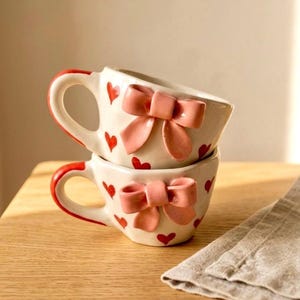 Puede incluir: Dos tazas de cerámica apiladas con una base de color crema, decoradas con pequeños patrones de corazones rojos. Cada taza presenta un lazo rosa claro tridimensional prominente en la parte frontal y un asa pintada de rojo en el lateral.