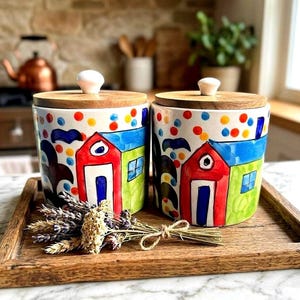 May include: Two ceramic canisters with wooden lids. The canisters are hand-painted with colorful houses, blue, red, and green, and polka dots. The canisters are on a wooden tray with dried flowers.